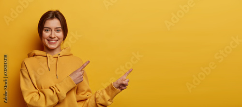 Check this out, better see it. Beautiful dark haired millennial girl wears casual hoodie, points both index fingers on blank space, has friendly smile, discusses promo, isolated on yellow background.