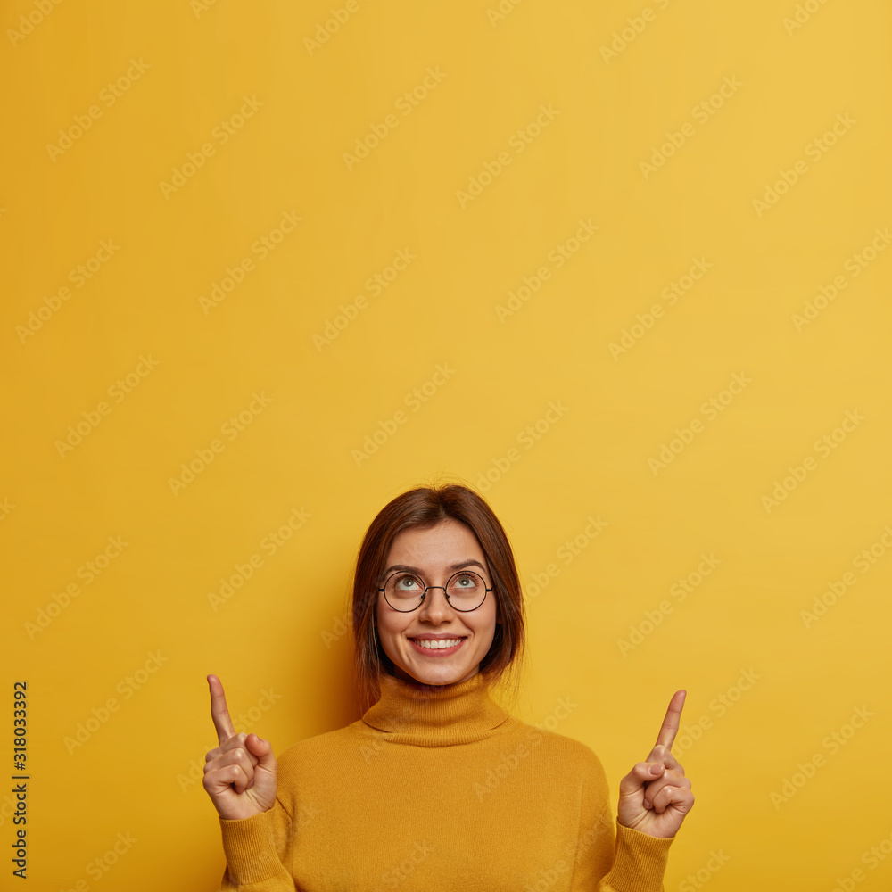Φωτογραφία Vertical image of attractive smiling woman points index fingers above, brags about her new ceiling, wears casual turtlenck, isolated over yellow background, has white perfect teeth