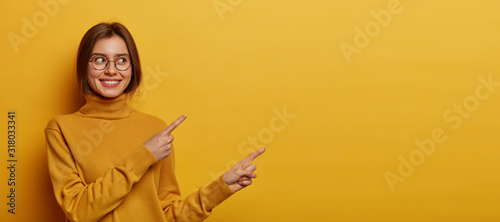 Studio shot of pleasant looking young woman with dark combed hair, wears neck sweater, points away on blank space, demonstrates advert, isolated over yellow background, describes amazing place