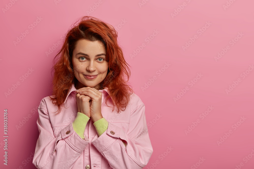 Φωτογραφία Portrait of redhead woman keeps hands under chin, happy to receive compliment, wears fashionable clothes, looks with positive emotions, has tender natural look, stands indoor over rosy wall