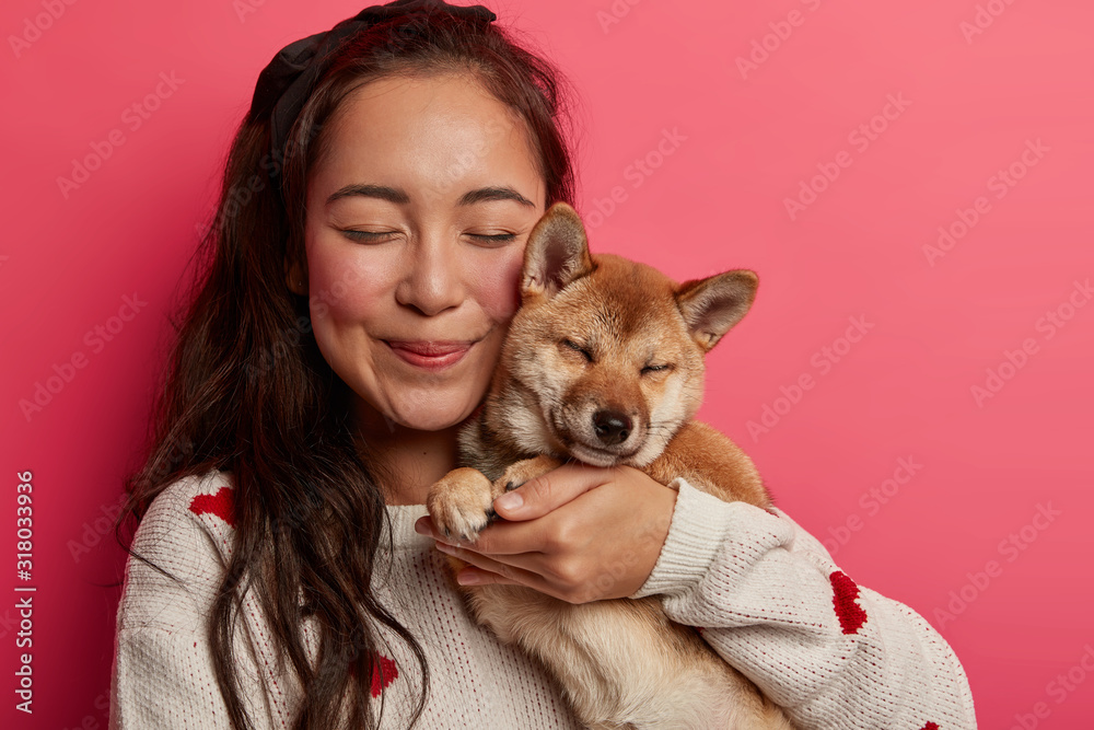 Close up shot of pleased Asian woman carries pedigree dog closely near  face, closes eyes from pleasure, embraces animal with love, makes memorable  shot with favourite pet, isolated over rosy wall Stock