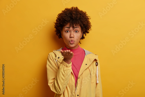 Romantic woman blows airkiss, keeps palms extended near mouth, sends mwah to boyfriend, has healthy skin, expresses her love, dressed casually, stands against vivid yellow background. Body language