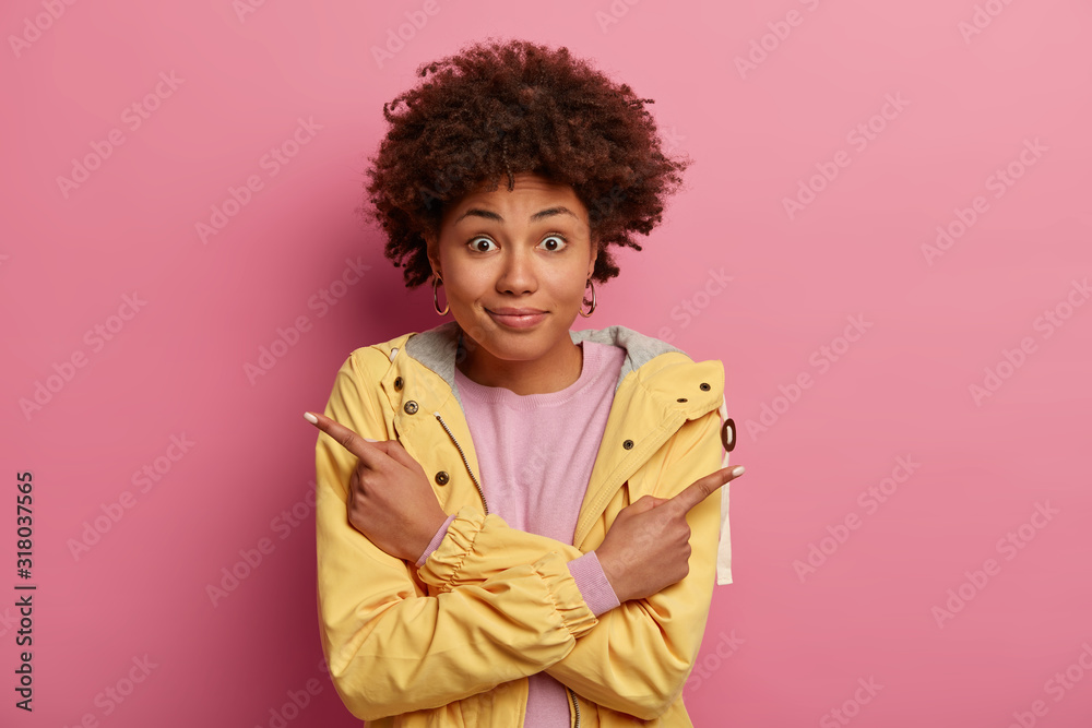 Confused positive woman with Afro hair points both index fingers in different sides, demonstrates two directions, shows something left and right, wears yellow clothes, crosses hands over chest