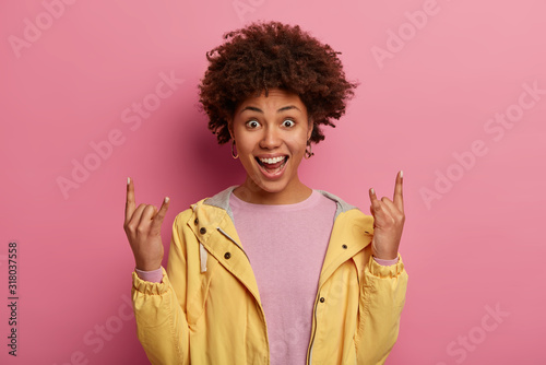 Charming overjoyed feminine girl makes rock n roll gesture, feels carefree and glad, listens favourite music, wears yellow jacket, feels upbeat isolated on rosy pastel wall. Hand sign. Rock this party