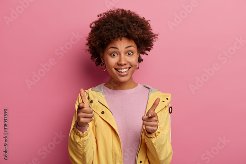You are chosen. Happy curly haired woman points index fingers at camera, says you are next, smiles broadly, expresses her choice, wears yellow anorak, makes approving gesture, isolated on pastel pink