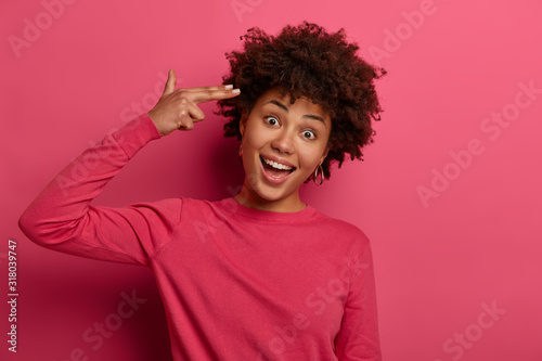 Funny glad young woman makes suicide gesture, finger gun, shoots in temple, tilts head, wears crimson jumper, isolated over pink background, commits suicide, has curly hairstyle. Monochrome shot
