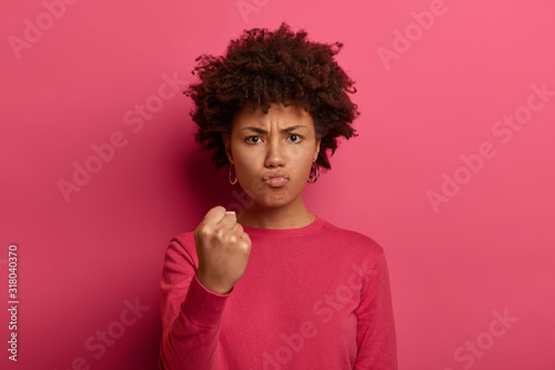 Irritated outraged young woman clenches fist, shows anger and looses temper, looks with annoyance at camera, expresses negative emotions, isolated over pink background, makes threaten gesture