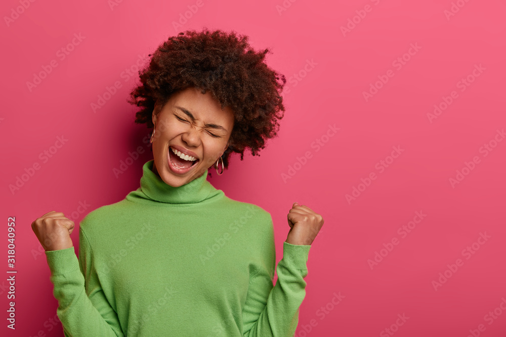 Great achievement and success concept. Happy Afro American woman clenches fists, makes victory gesture, screams yeah, tilts head, wears green turtleneck, celebrates achieving goal, isolated on pink Great achievement and success concept. Happy Afro American woman clenches fists, makes victory gesture, screams yeah, tilts head, wears green turtleneck, celebrates achieving goal, isolated on pink