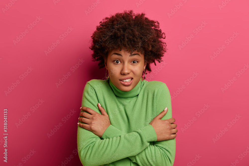 How chilly. Puzzled dark skinned lady hugs herself, trembles from cold, walks on windy weather, shakes and clenches teeth, dressed in green sweater, stands without coat, isolated on pink wall