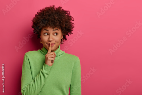 Pretty woman makes shush, asks not be loud, has secret expression, presses index finger over lips, has mysterious expression, wears green jumper, isolated over rosy wall with free space, keeps quiet photo