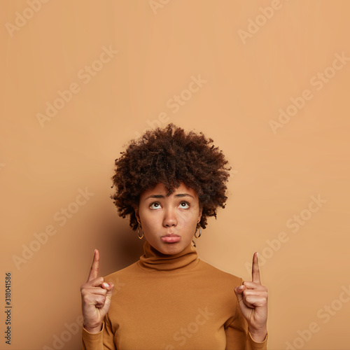 Unhappy curly haired woman dislikes something, points above, purses lower lip, has gloomy face expression, wears poloneck, concentrated upwards sadly, isolated over beige background. Monochrome