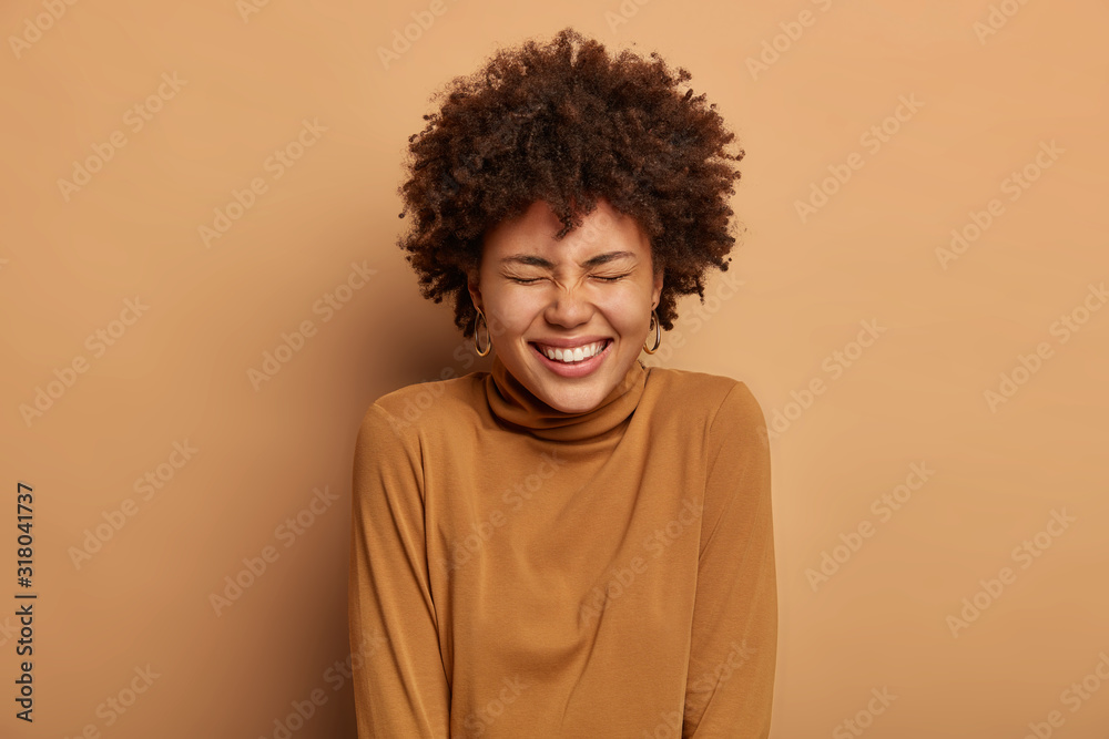 Portrait of joyous glad lady with toothy smile, laughs out as hears comic story, being in high spirit, enjoys happy day, has natural curly hair, isolated over beige background. Upbeat emotions Portrait of joyous glad lady with toothy smile, laughs out as hears comic story, being in high spirit, enjoys happy day, has natural curly hair, isolated over beige background. Upbeat emotions