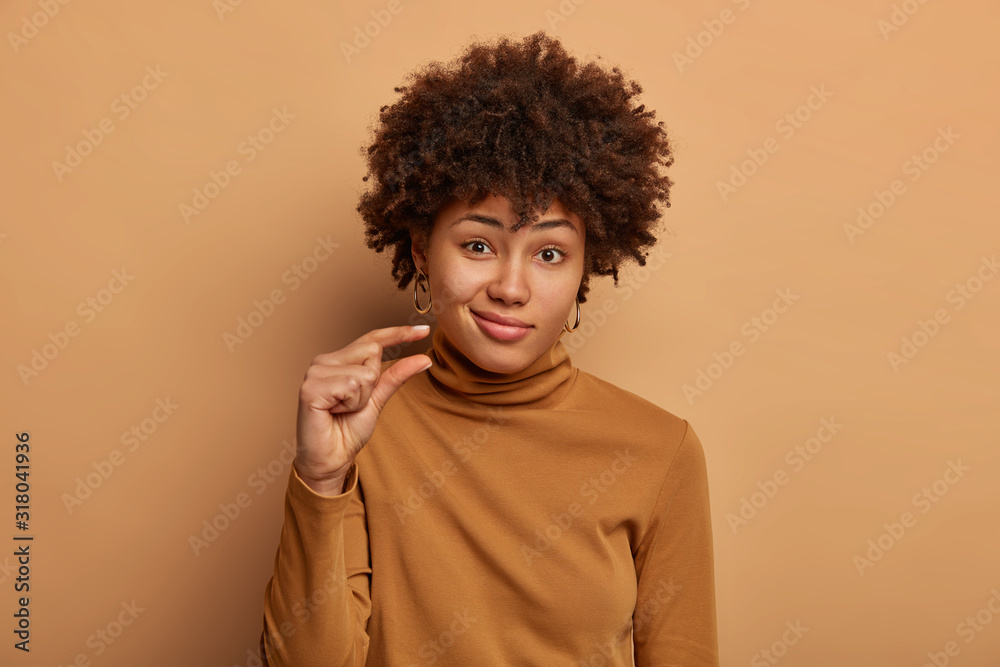 Portrait of attractive woman with curly Afro hair, shapes tiny and small thing, talks about size, dressed in casual brown jumper, being unimpressed, isolated over beige studio wall. Little object