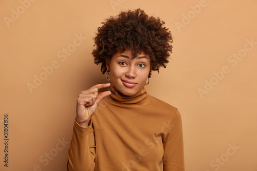 Portrait of attractive woman with curly Afro hair, shapes tiny and small thing, talks about size, dressed in casual brown jumper, being unimpressed, isolated over beige studio wall. Little object © wayhome.studio 