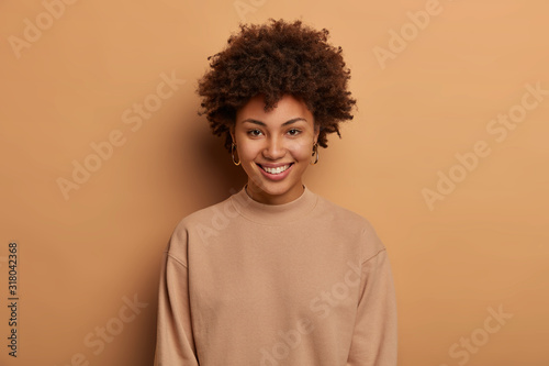 Modest relaxed healthy Afro American woman has tender toothy smile, enjoys lovely day, expresses positive emotions, wears casual brown jumper, looks directly at camera with eyes full of happiness. © wayhome.studio 