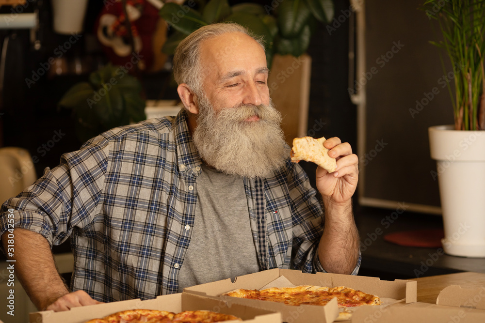 Smiling older man eating pizza slice sitting at living room. Bearded senior man eating italian food. Smiling older man eating pizza slice sitting at living room. Bearded senior man eating italian food.