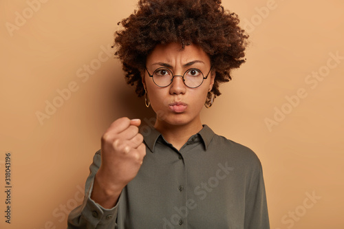 Outraged angry woman shows clenched fist to camera, has strict facial expression, needs respect, demands good behaviour, expresses anger and irritation, threatens to revenge, wears stylish shirt © wayhome.studio 