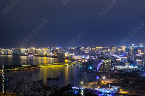 Night view of Sanya city with bright multi-colored illumination buildings, structures, roads, sidewalks, poles, bridges.