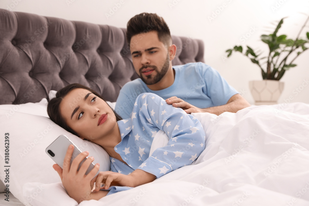 Fototapeta premium Young woman ignoring her distrustful boyfriend while using smartphone in bed at home