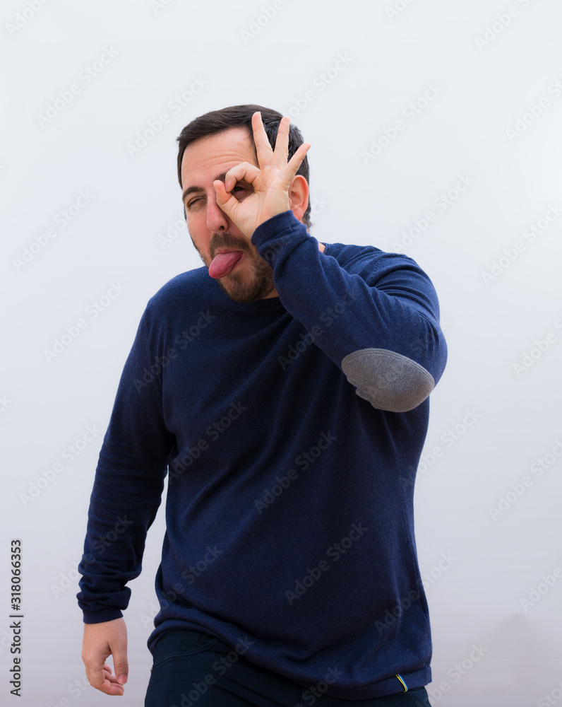 young handsome man smiling happily with funny face, joking and looking through peephole, spying on secrets against white wall. young handsome man smiling happily with funny face, joking and looking through peephole, spying on secrets against white wall.