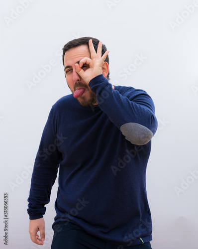 young handsome man smiling happily with funny face, joking and looking through peephole, spying on secrets against white wall.