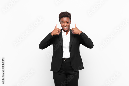 young pretty black womansmiling broadly looking happy, positive, confident and successful, with both thumbs up against white wall photo