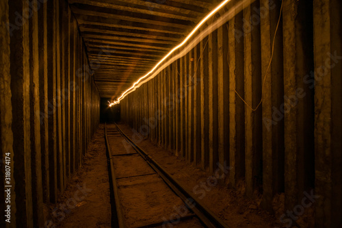 Fototapeta Naklejka Na Ścianę i Meble -  Underground square tunnel with rails for minecart and warm lighttrail.