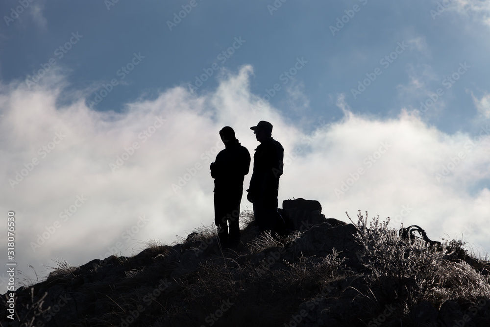 Silhouettes of two mountain hikers standing on the peak covered by frozen grass against a background blue sky