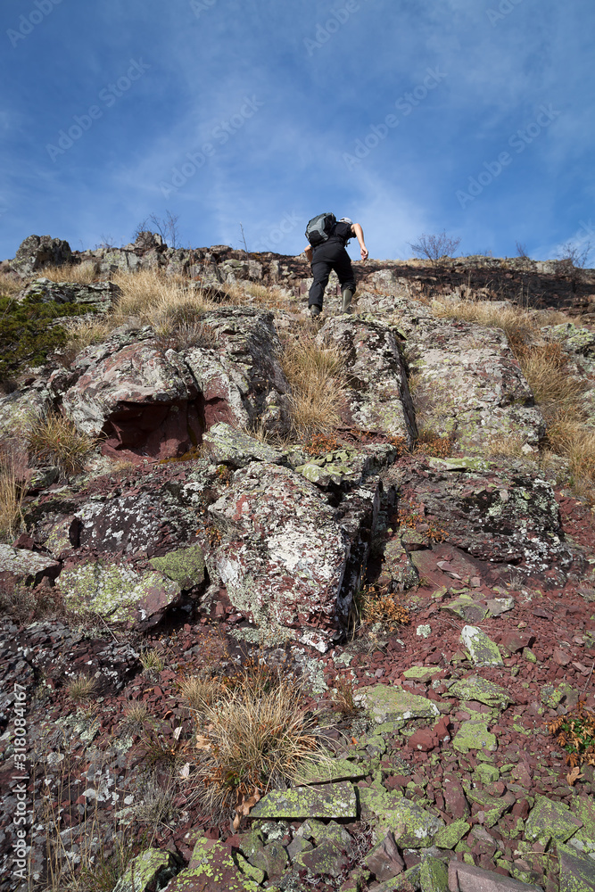 Mountain hiker with backpack and rubber boots, dressed in black, climbing up the steep, rocky, dangerous cliff covered by red and green rocks and ground burned by fire