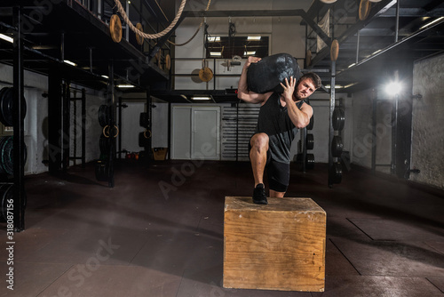 Young strong sweaty fit muscular man with big muscles holding heavy sandbag on his shoulder with his hands and climb on the jump box for legs cross workout training in the gym photo