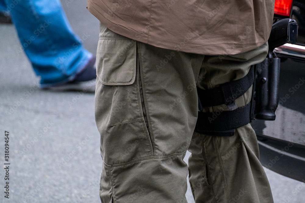 A police office standing on pavement with a gun strapped to his thigh or right leg. The cop is wearing green cargo pants with large pockets and a tan color coat.  There are people in the background.