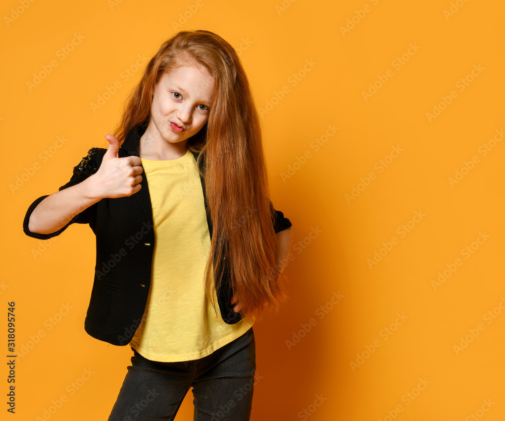 Red-haired schoolgirl in black jacket and pants, yellow t-shirt. She is showing thumb up, posing on orange background. Close up Red-haired schoolgirl in black jacket and pants, yellow t-shirt. She is showing thumb up, posing on orange background. Close up