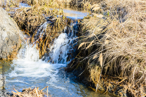 Long-awaited spring creeks flow over ravines and hills on a sunny day. Water rapids and waterfalls of streams among the dry grass. Spring landscape. © Evgeniy