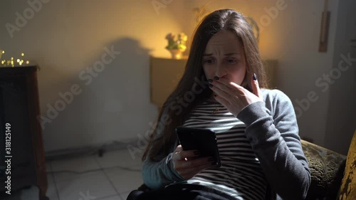 Scared girl sitting on armchair and looking on cellphone display in a dusk room. Girl receiving horror messages and showing her emotions offear. Horror atmosphere in dark room, retro style furniture photo