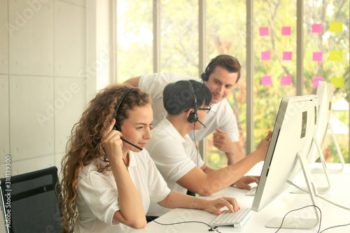 Young beautiful woman with headphones working at call center service desk consultant with her teammates while team supervisor helping new teammate and training junior to talk with the customer on hand
