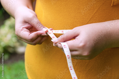Hand fat woman holding measuring tape around her waist. Overweight female measuring her waist with measuring tape while standing in the garden. Healthcare concept.
