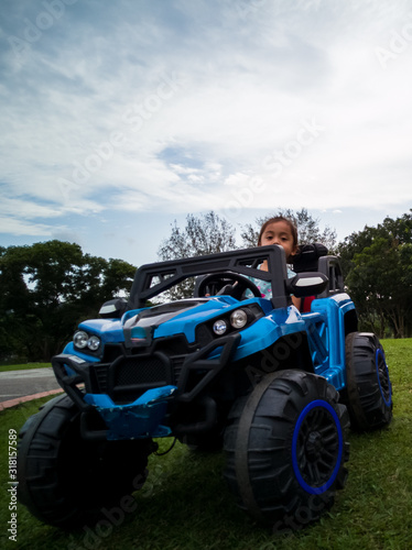 An Asian kid driving electric toy car in a tropical park. Outdoor toys. Children in battery power vehicle. Little girl riding toy truck in the garden. Family playing in the backyard.