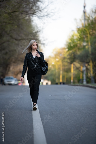 A young beautiful blonde girl in a black leather jacket is walking along an empty city road, the lower angle of the shooting