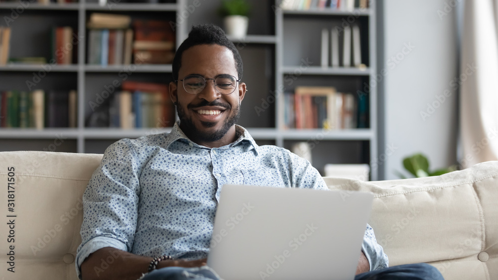 Smiling african man using laptop watching movies sit on sofa - obrazy, fototapety, plakaty 