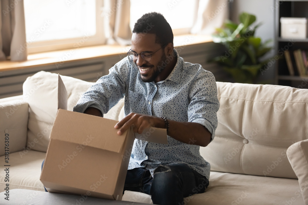 Smiling african man customer opening cardboard box parcel on sofa Smiling african man customer opening cardboard box parcel on sofa