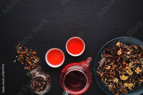 Aromatic fresh brew natural red fruit tea in two bowls ,glass teapot,glass jar and plate with dry tea leaves on a black concrete background