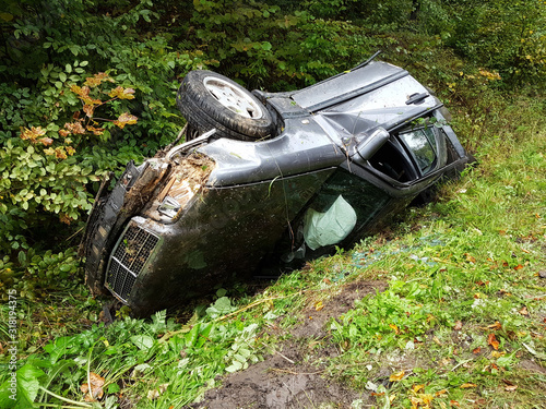 A battered gray car sedan lies in a roadside ditch among the lush green foliage of the bush in the summer. Road traffic incident. The consequences of speeding and the threat of life. Traffic safety