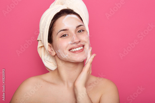 Charming female after applying cream on face, young girl keeping hand on her cheek, posing isolated over pink studio background, beautiful woman with bare shoulders doing beauty procedures at home.