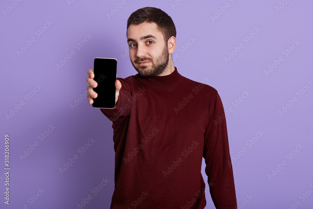 Photo of young handsome bearded guy wearing dark burgundy sweater, showing mobile phone with blank screen at camera isolated over lilac background. Copy space foer advertisment or promotional text. Photo of young handsome bearded guy wearing dark burgundy sweater, showing mobile phone with blank screen at camera isolated over lilac background. Copy space foer advertisment or promotional text.