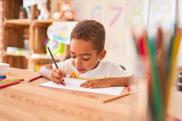 Beautiful african american toddler sitting drawing using paper and pencils on desk at kindergarten