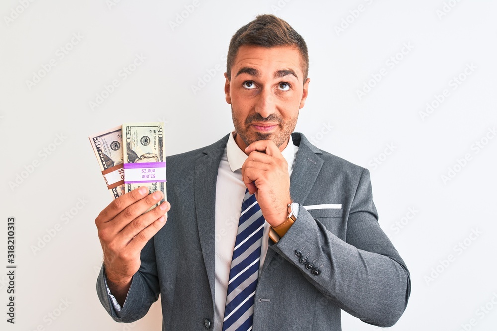 Young handsome business man holding bunch of dollars banknotes over isolated background serious face thinking about question, very confused idea Young handsome business man holding bunch of dollars banknotes over isolated background serious face thinking about question, very confused idea