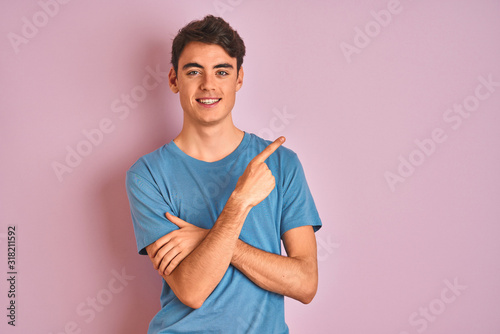 Teenager boy wearing casual t-shirt standing over blue isolated background with a big smile on face, pointing with hand and finger to the side looking at the camera.