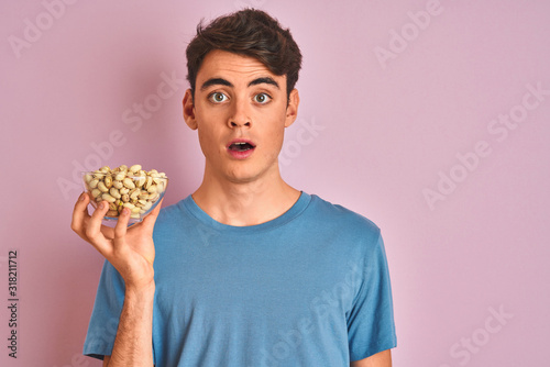 Teenager boy holding a bunch of pistachios over isolated pink background scared in shock with a surprise face  afraid and excited with fear expression
