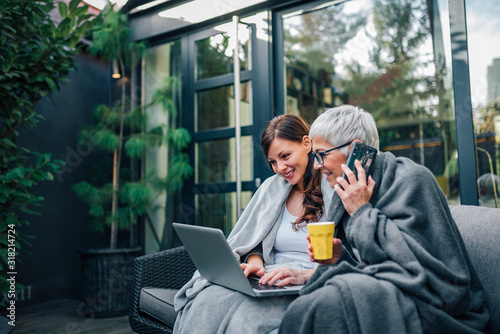 Two successful women of different age sitting in the garden, in blankets, using laptop and talking on smart phone.