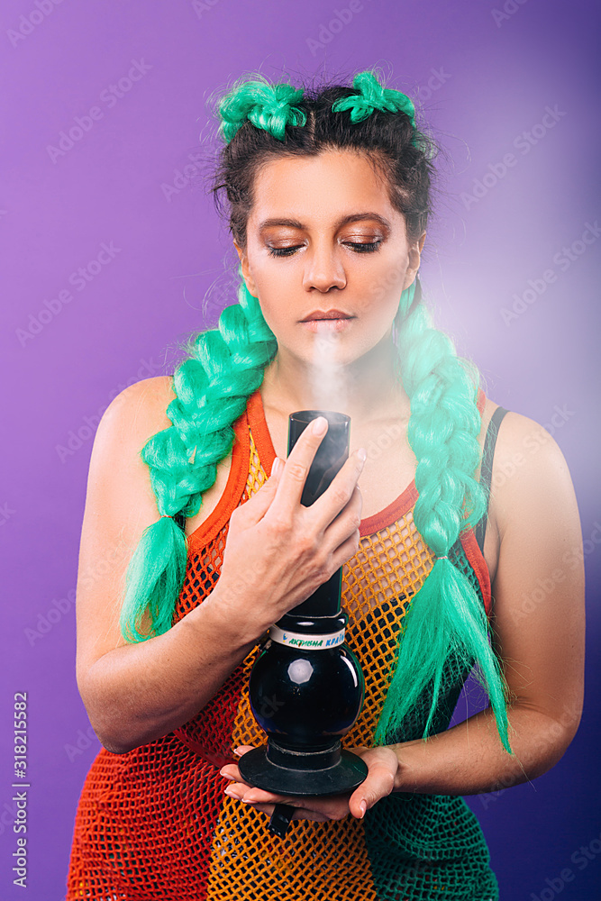 Big glass bong in the hands of a hipster woman. Woman smokes cannabis. Female hairstyle with green kanekalon on violet background Big glass bong in the hands of a hipster woman. Woman smokes cannabis. Female hairstyle with green kanekalon on violet background